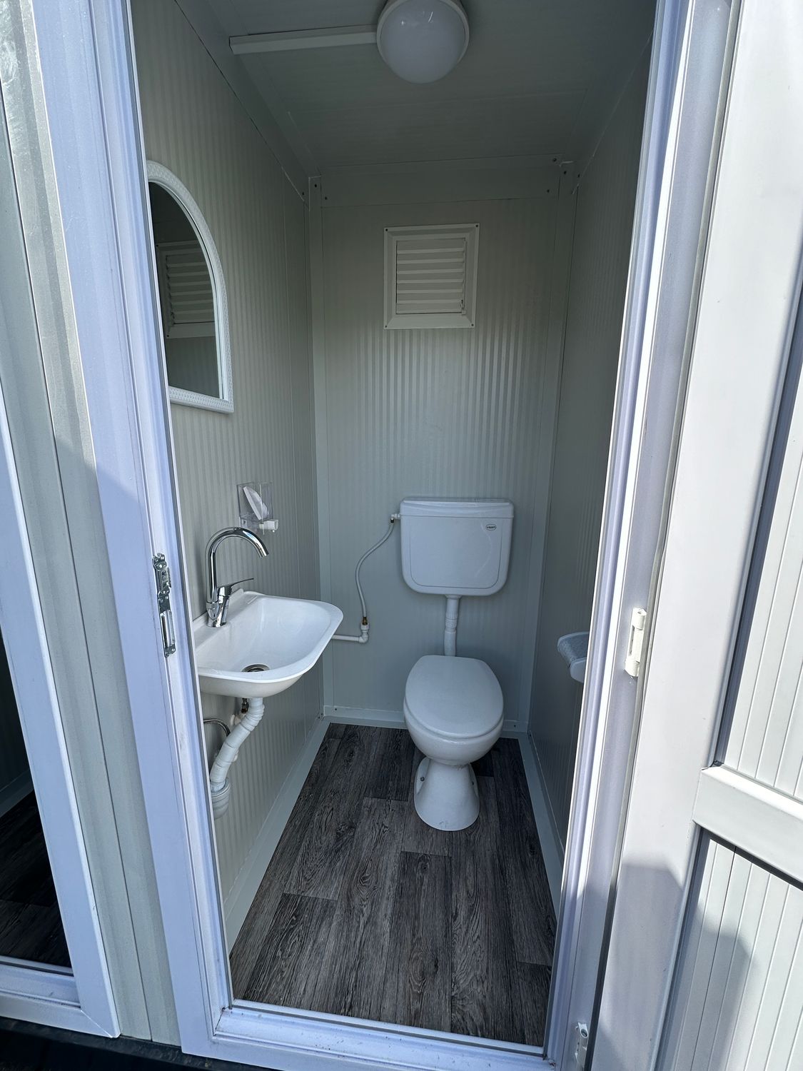 Blick in eine WC-Kabine mit Waschbecken, Spiegel, Lüftungsgitter und dunklem Holzdekorboden