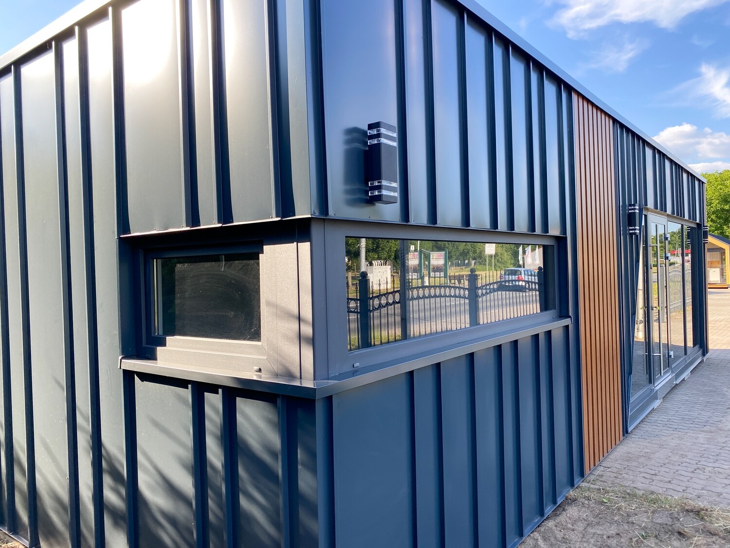 Schrägansicht der dunkelblauen Fassade mit Fensterband, aufgestellter Klappe, Wandleuchte und Holzstreifen