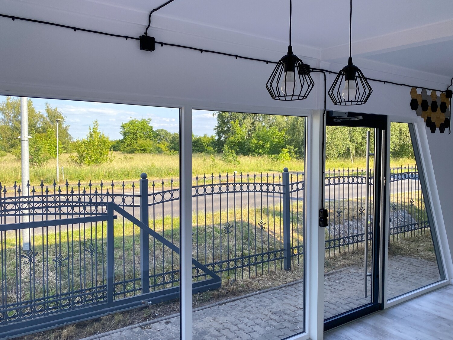 Fensterfront von innen mit Blick auf Zaun und Wiese, darüber Kabelleuchten an der Decke