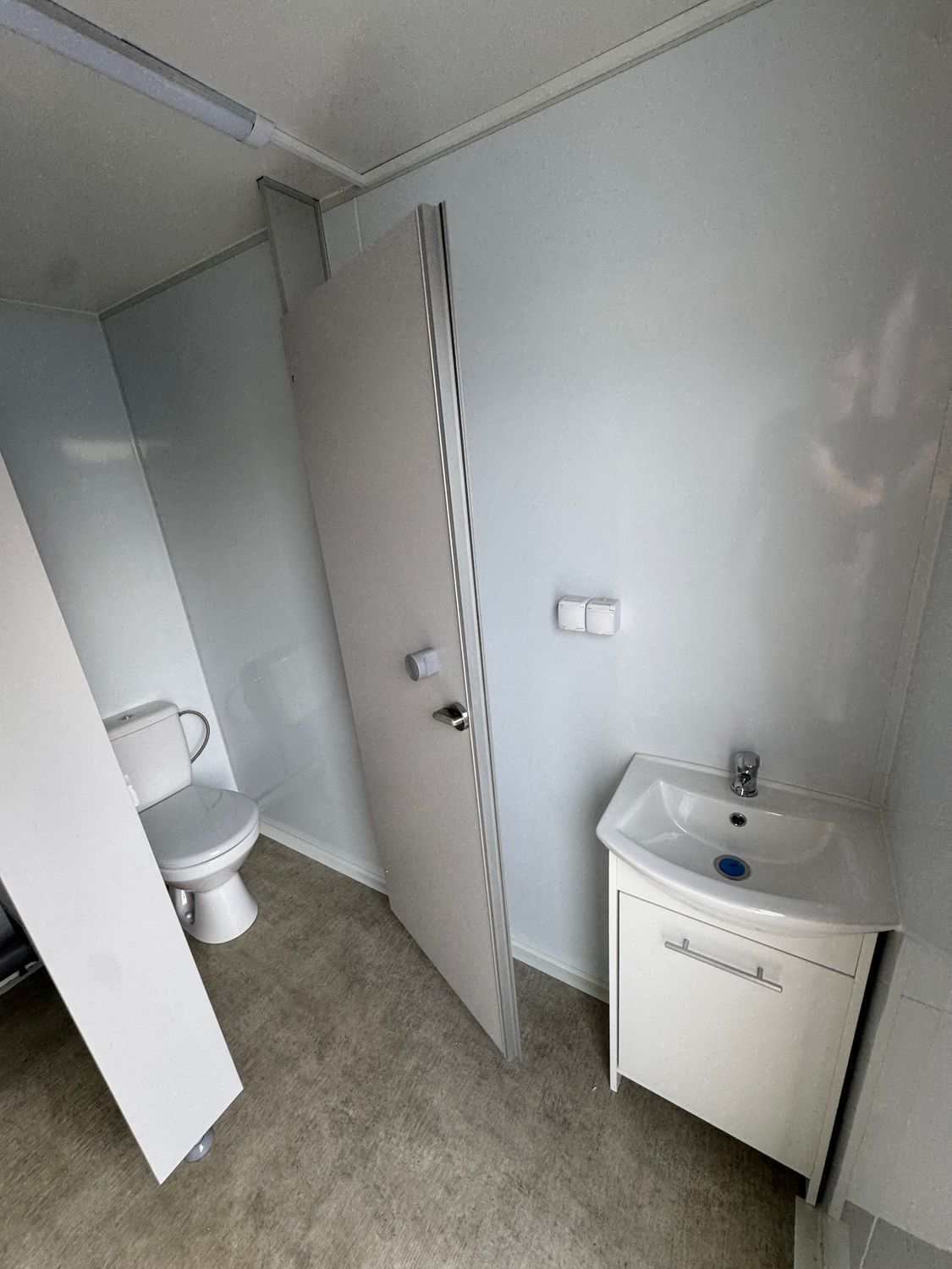 Innenansicht mit offener Kabinentür und WC dahinter, rechts Waschtisch mit Unterschrank und Schaltern