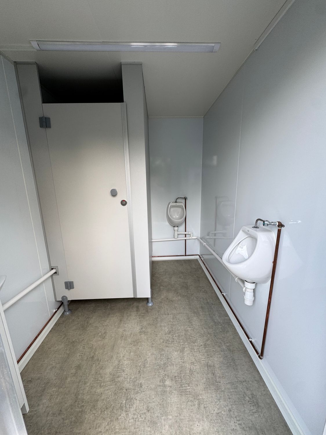 Herrenbereich mit zwei wandhängenden Urinalen, sichtbaren Leitungen und geschlossener Kabinentür