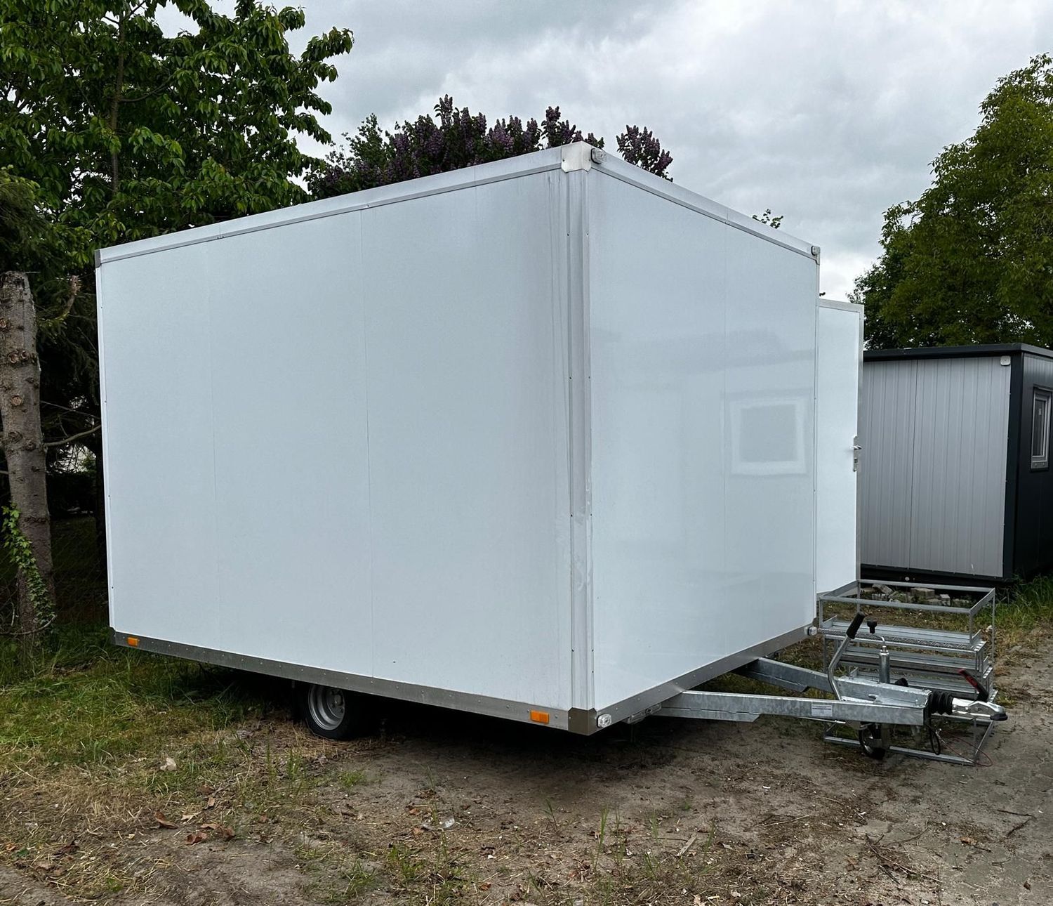 Stirnseite des weißen WC-Anhängers mit Deichsel, Stützrad und Einachsfahrwerk auf einer Wiese