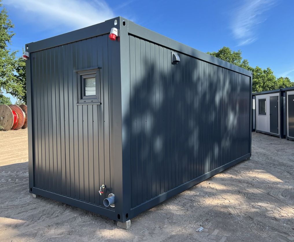 Schräg von hinten fotografierter Wohncontainer mit kleinem Fenster, roten CEE-Anschlüssen und Abflussrohr