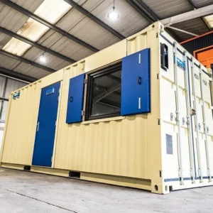 Beiger 20-Fuß-Bürocontainer in einer Halle, blaue Tür und Fenster mit aufgeklappten Rollladenflügeln