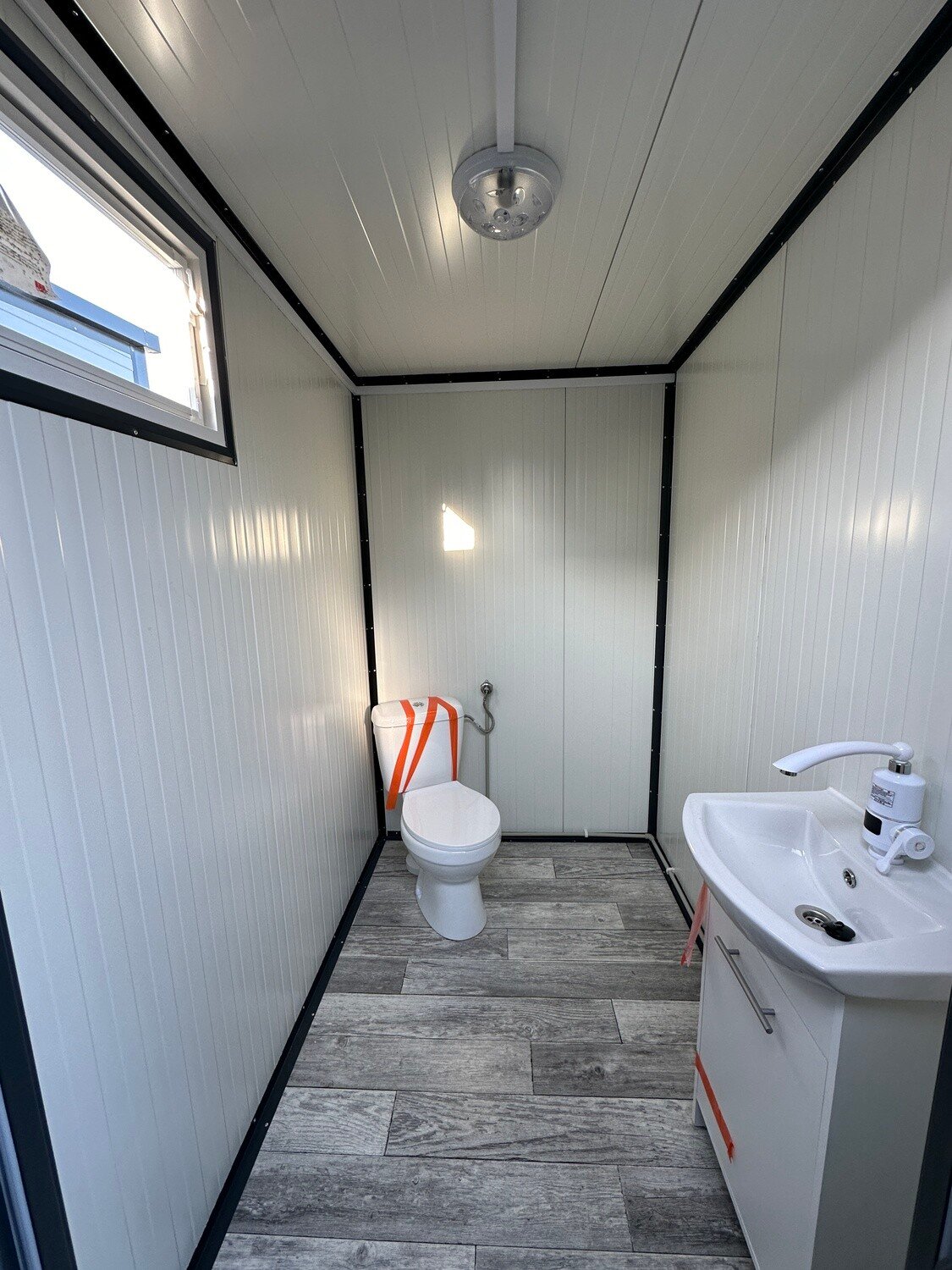 Innenansicht einer Kabine mit Toilette, Waschtisch mit Unterschrank und Deckenleuchte im Doppel-WC Container