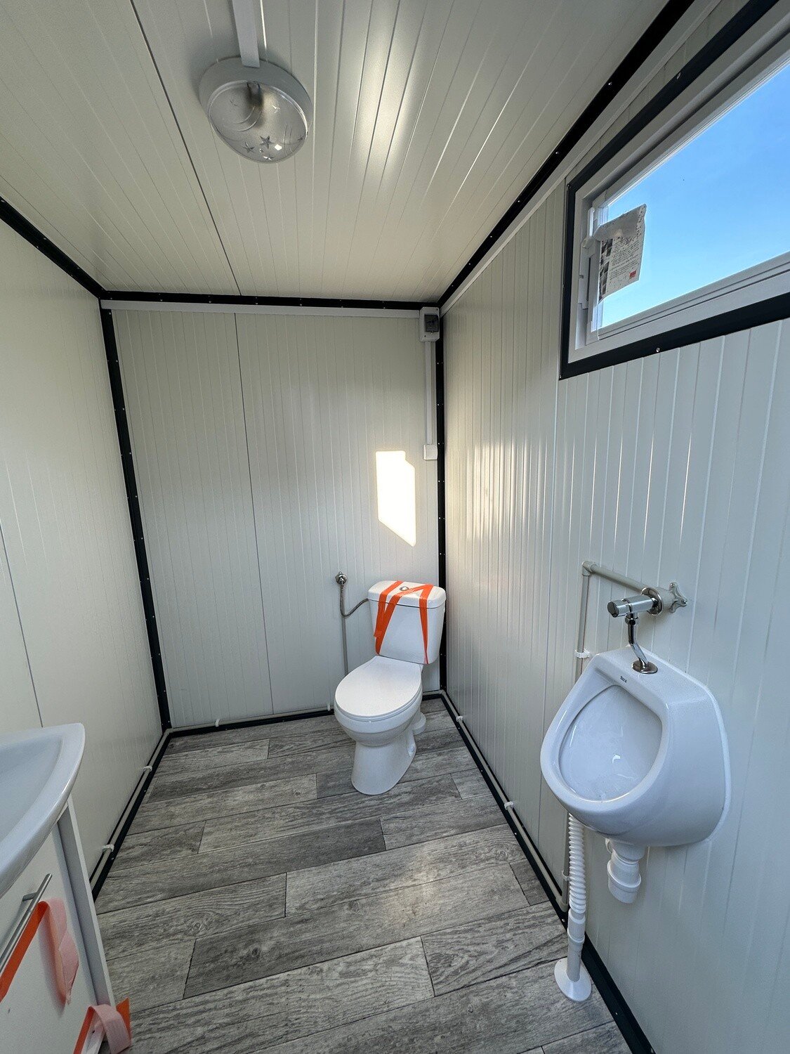 Zweite Kabine des Doppel-WC Containers mit Toilette, Urinal und Fenster über Bodenbelag in Holzoptik