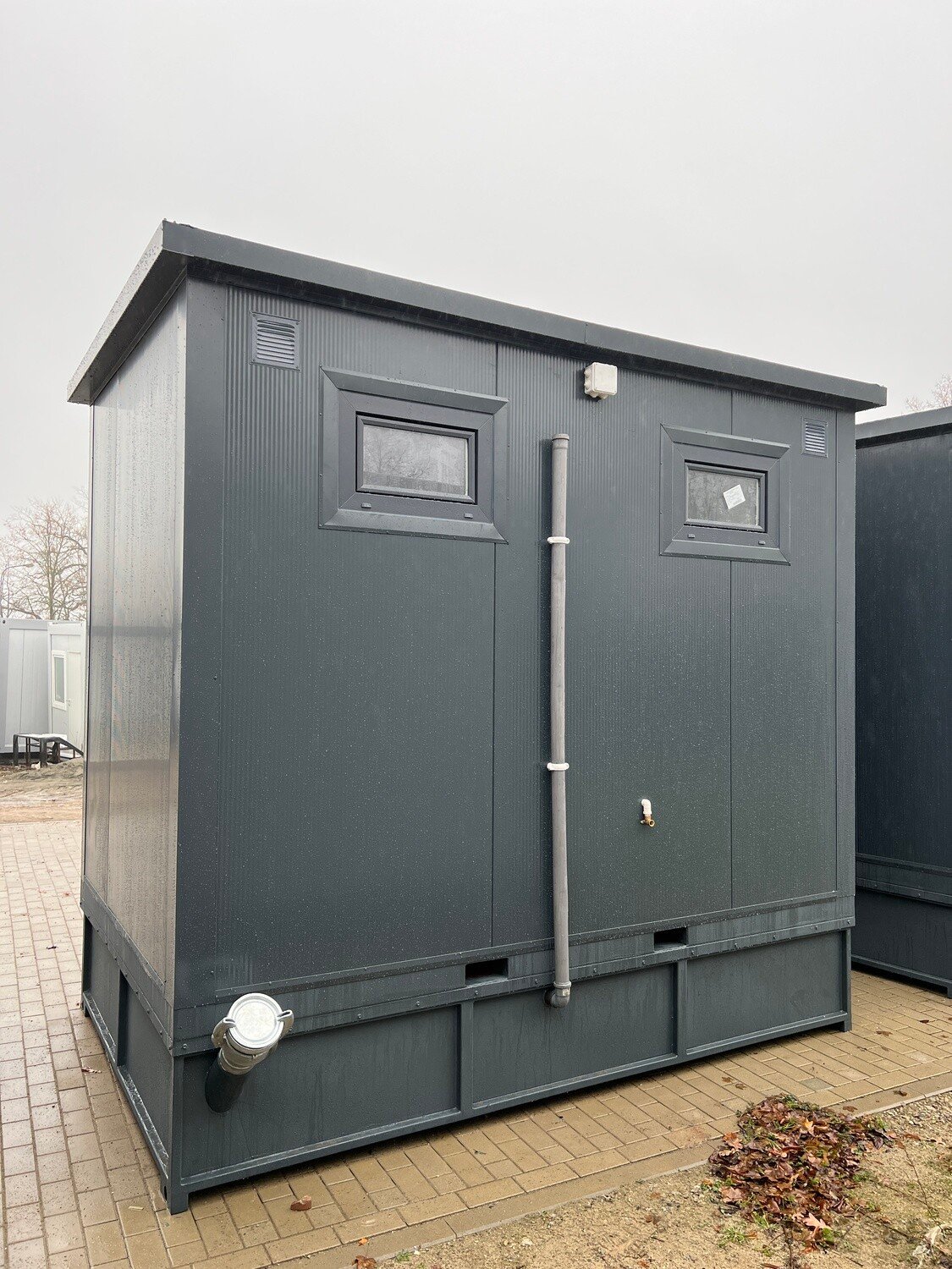 Rückansicht des WC Containers mit zwei kleinen Fenstern, senkrechtem Rohr und Anschlussstutzen am Sockel