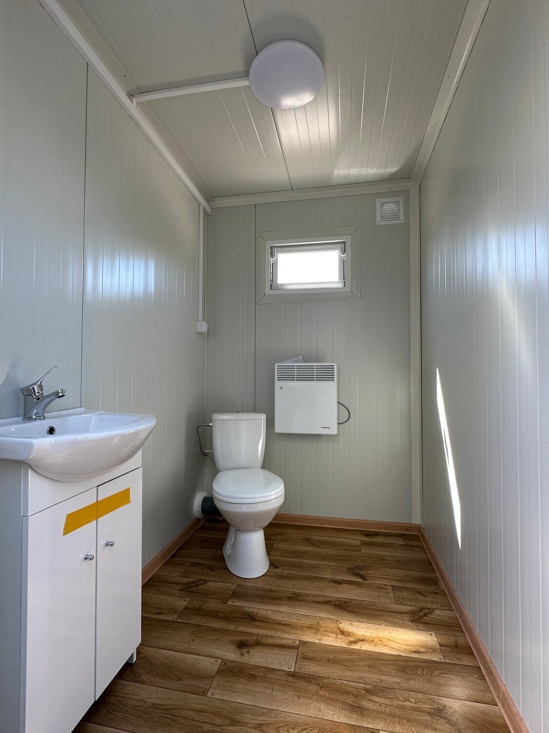 Helle Sanitärkabine mit Waschtisch, WC, Wandheizung und Fenster, Deckenleuchte über dem Holzoptik-Boden