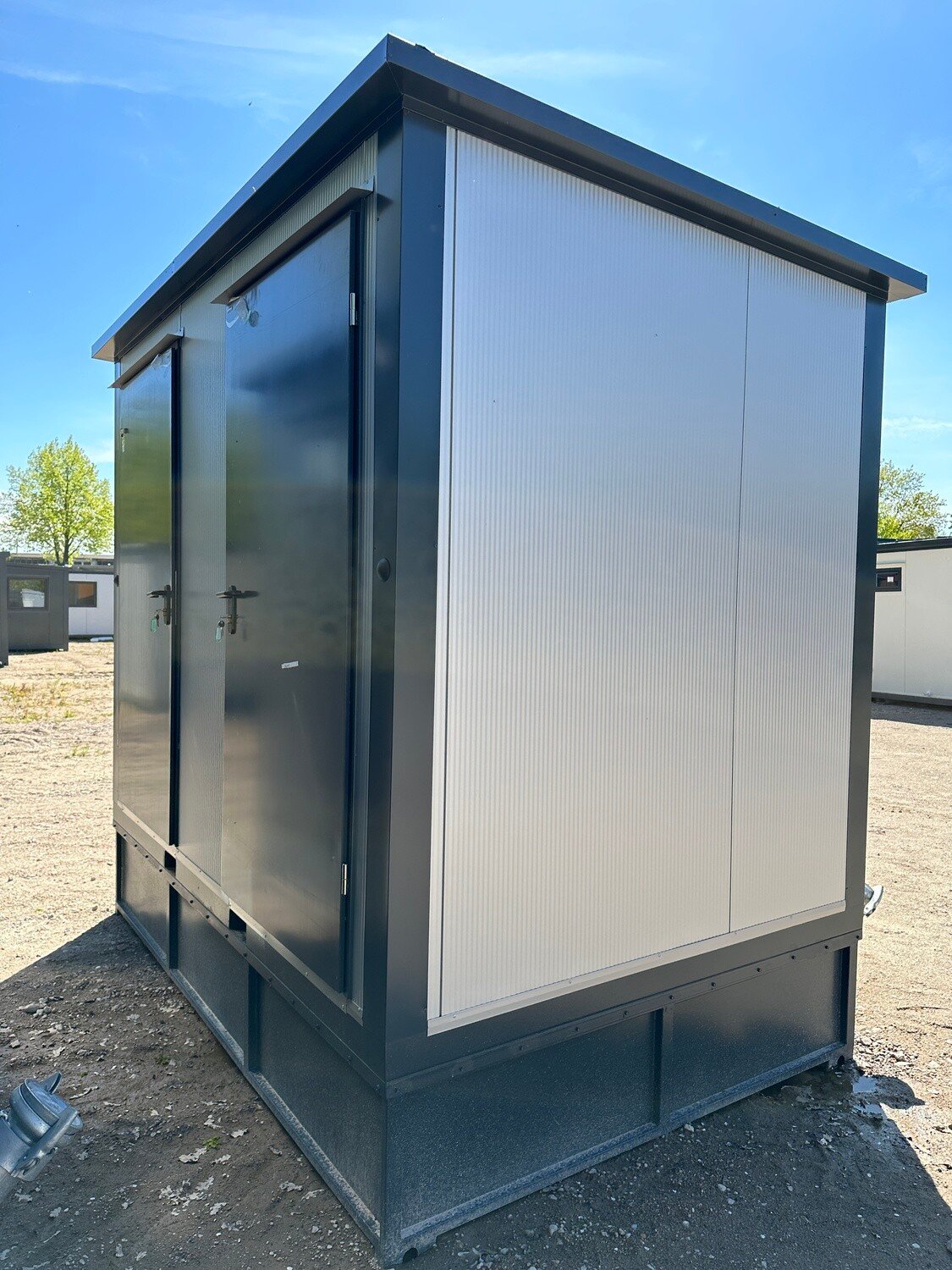 Doppel-WC Container mit hellen Wandpaneelen und dunklem Rahmen, zwei Türen mit Griffen auf Kiesfläche