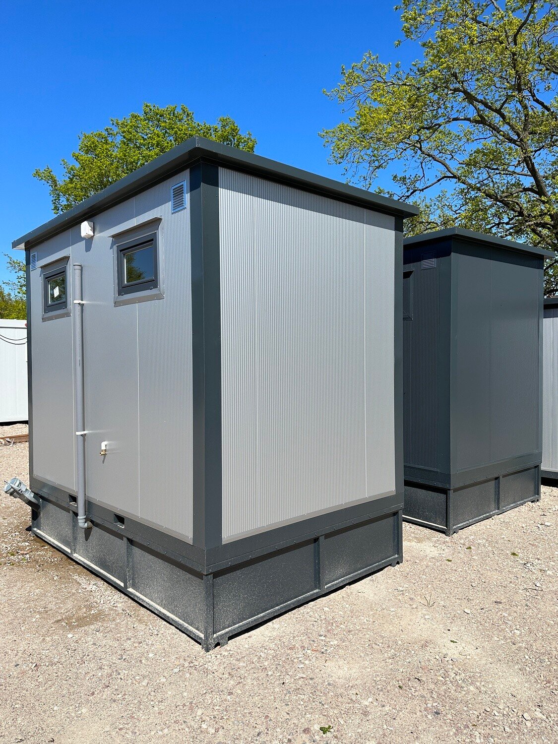 Seitenansicht eines grauen WC Containers mit kleinen Fenstern und Fallrohr, dahinter ein zweiter Container