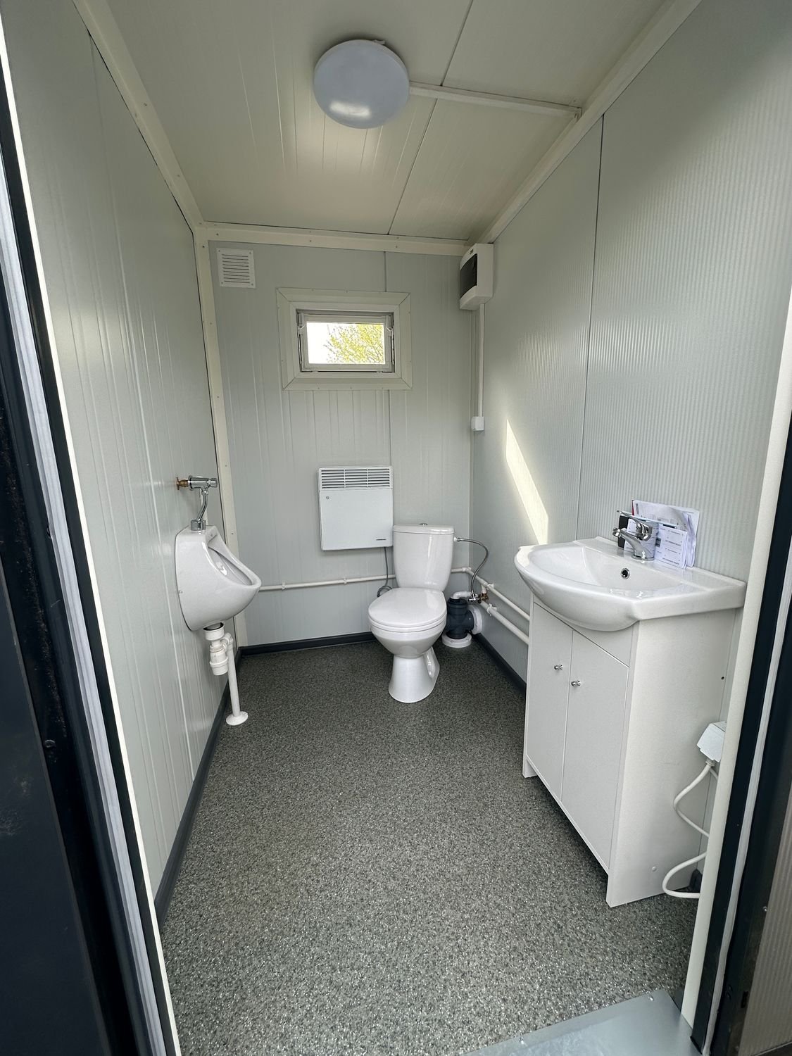 Innenansicht einer Kabine mit Urinal, WC, Wandheizung, Waschbecken auf Unterschrank und gesprenkeltem Boden