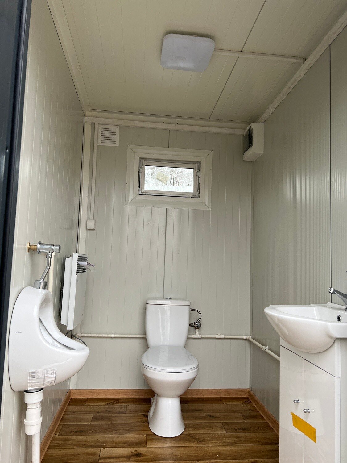 Kabine mit Urinal und WC über holzoptischem Bodenbelag, rechts Waschtisch mit weißem Unterschrank