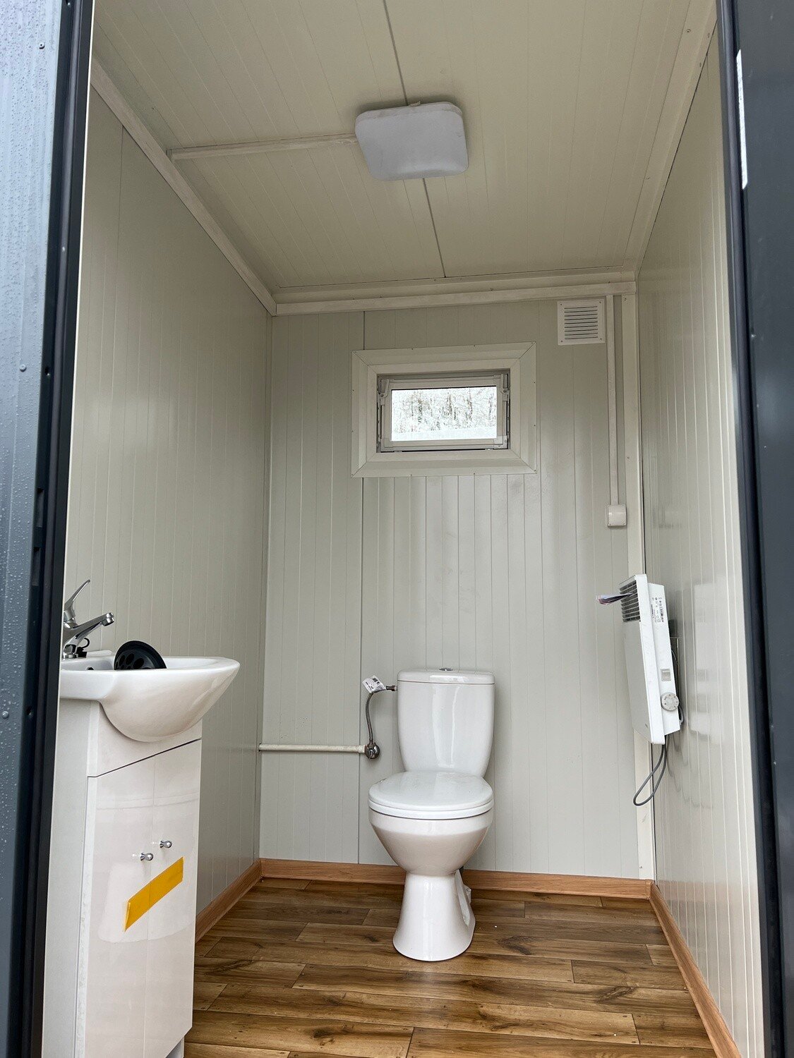 Blick in die WC-Kabine mit Waschbecken, Toilette, Heizgerät an der Wand und Boden in Holzoptik