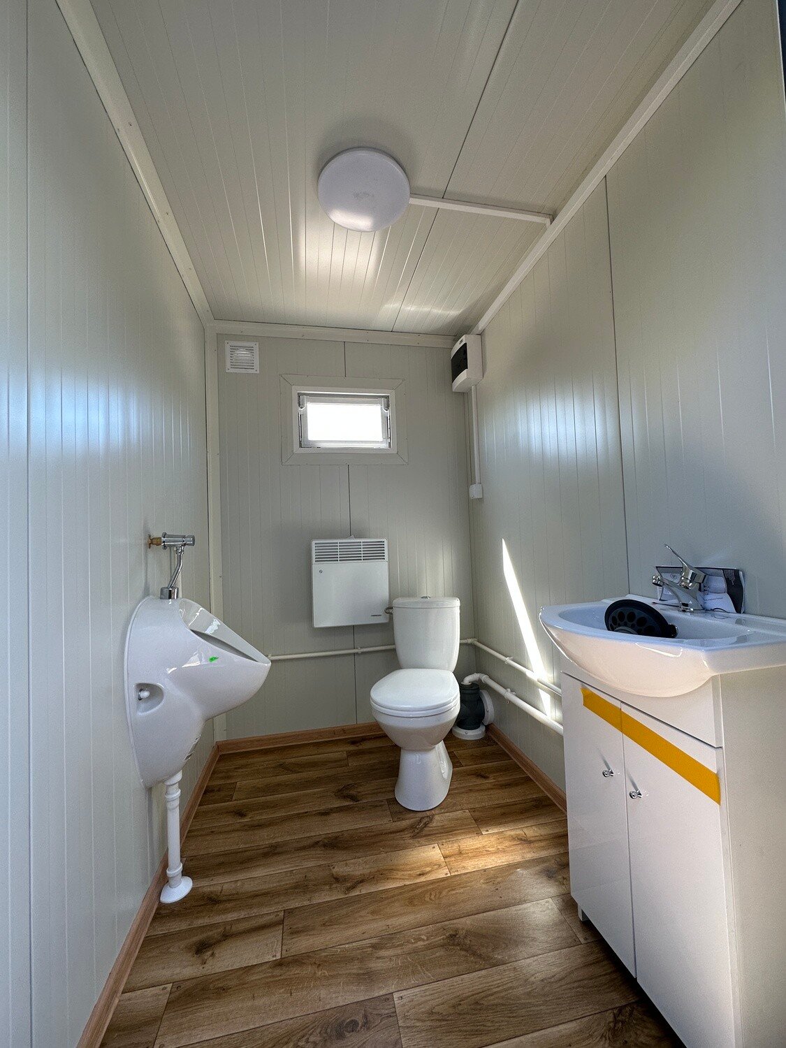 Innenraum mit Urinal, Toilette, Heizung unter dem Fenster und Waschtisch, darunter Bodenbelag in Holzoptik