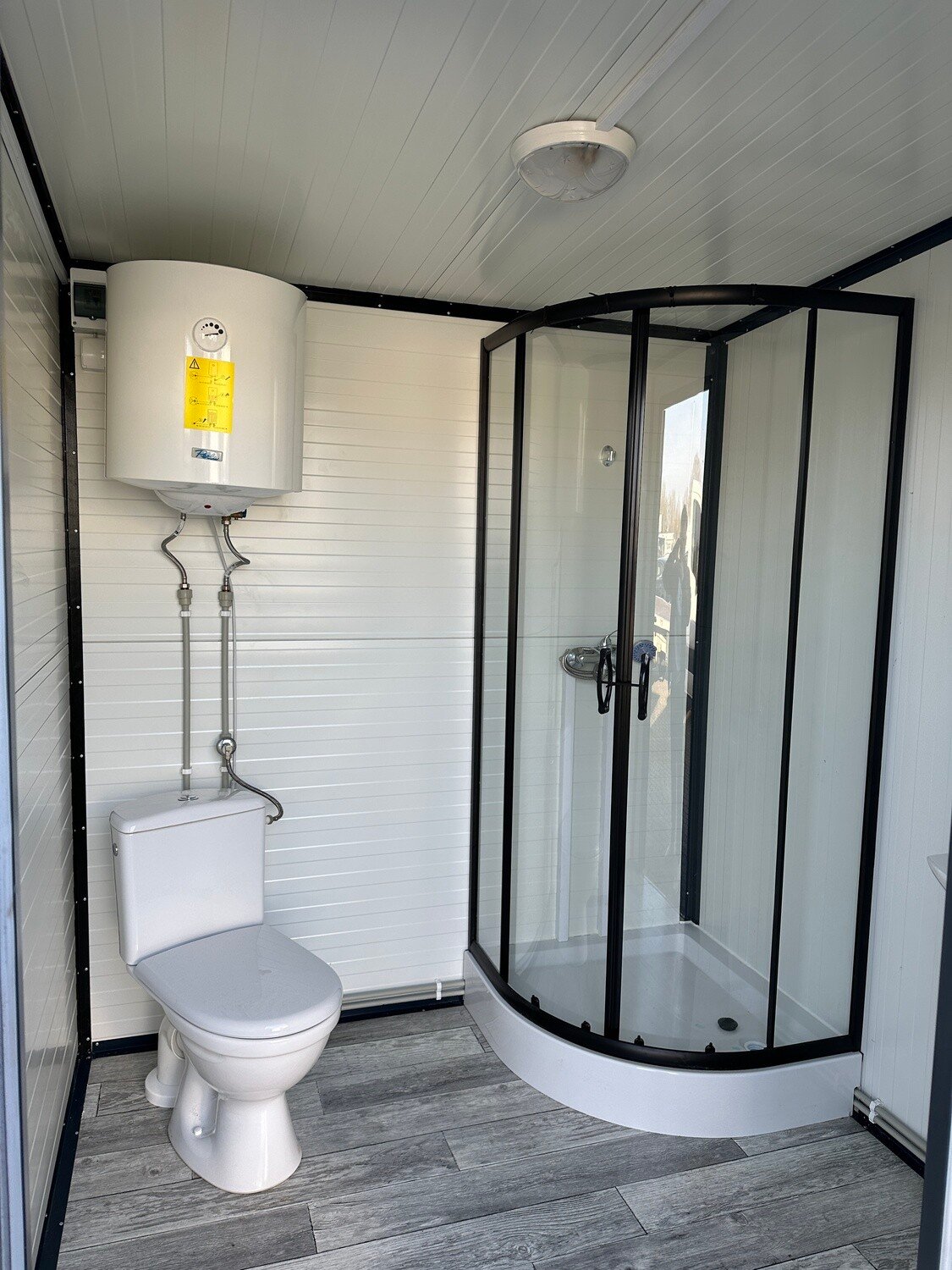 Innenansicht: Warmwasserboiler über dem WC, rechts daneben die Duschkabine mit dunklem Rahmen