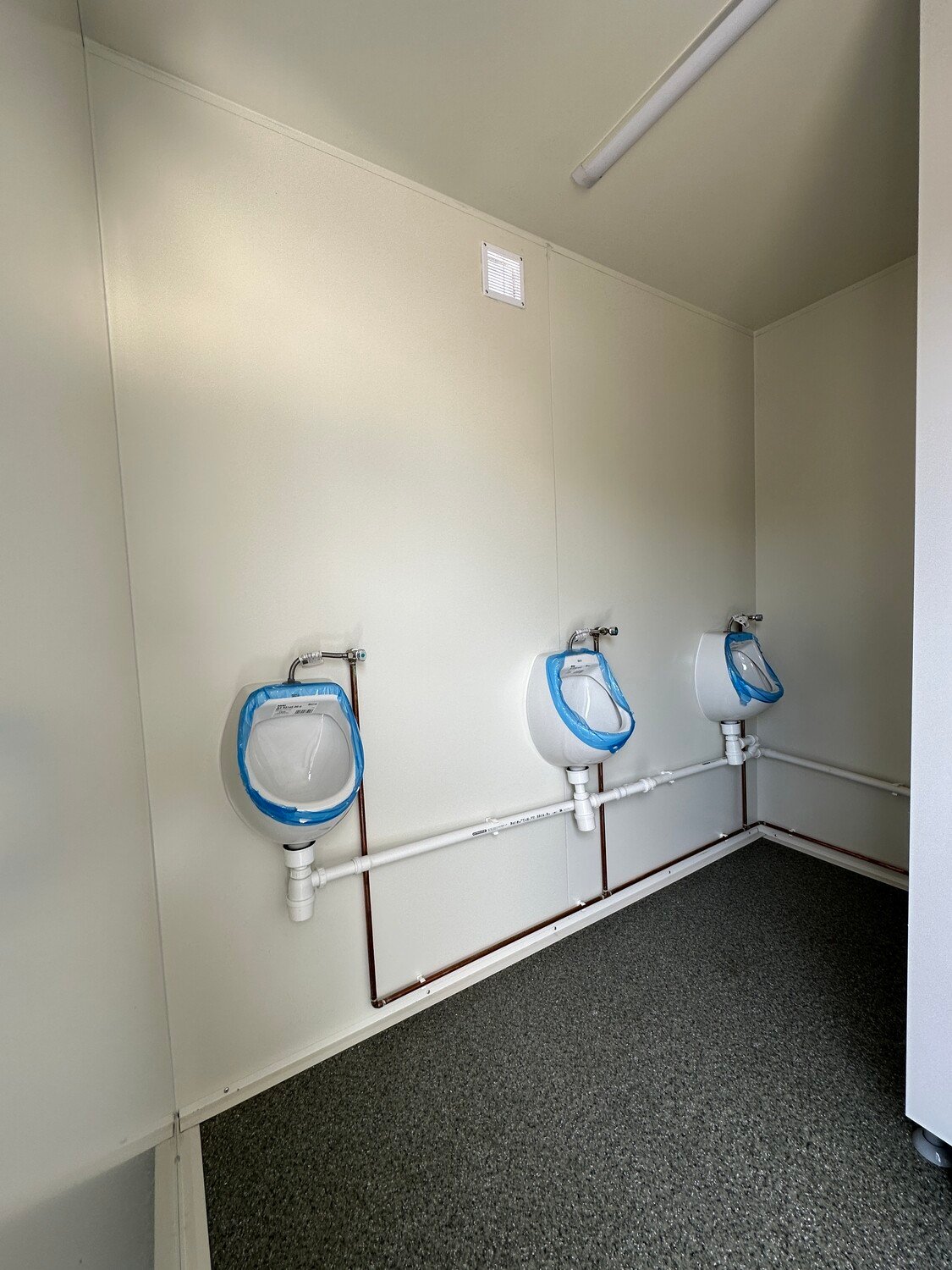 Innenraum des WC-Anhängers mit drei wandhängenden Urinalen, Lüftungsgitter und grauem Bodenbelag
