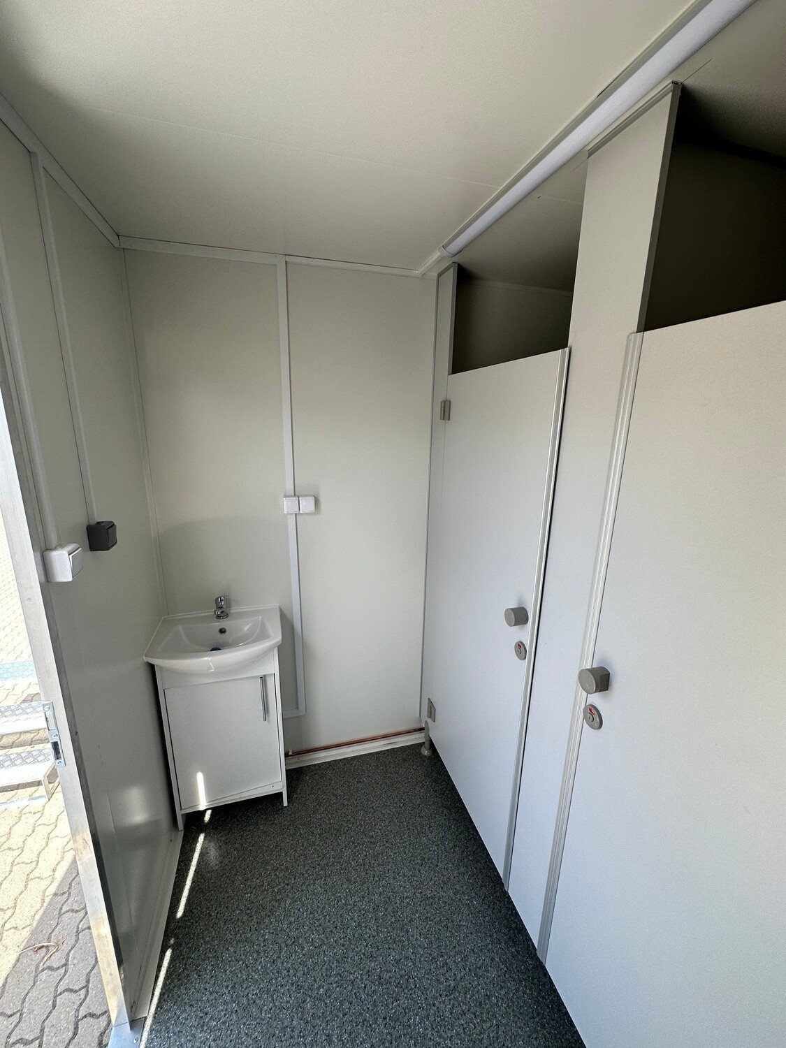 Blick durch die offene Tür auf Waschbecken mit Unterschrank, Seifenspender und zwei Kabinentüren