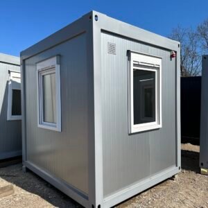 Hellgrauer Bürocontainer 3,00 x 2,44 auf Kiesfläche, mit zwei Fenstern, Lüftungsgitter und Stapelecken