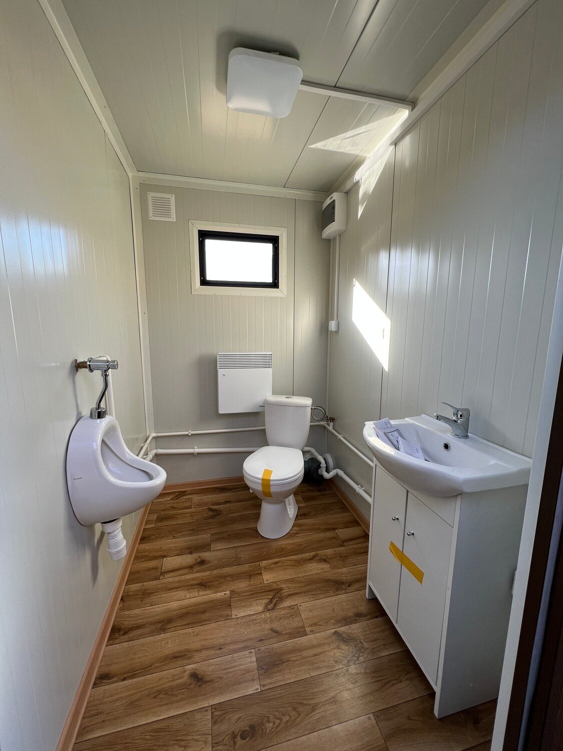 Innenansicht mit Urinal, WC, Waschtisch und Wandheizkörper über Holzdekor-Boden im Doppel-WC Container
