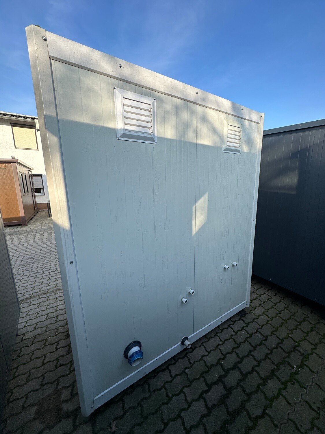 Weiße Außenwand des Sanitärcontainers 1,30 x 2,05 m mit zwei Lüftungsgittern und Rohranschlüssen