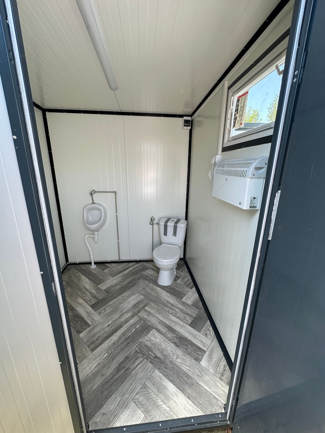 Blick durch die geöffnete Tür auf WC, Urinal, Fenster und Wandheizgerät des Sanitärcontainers
